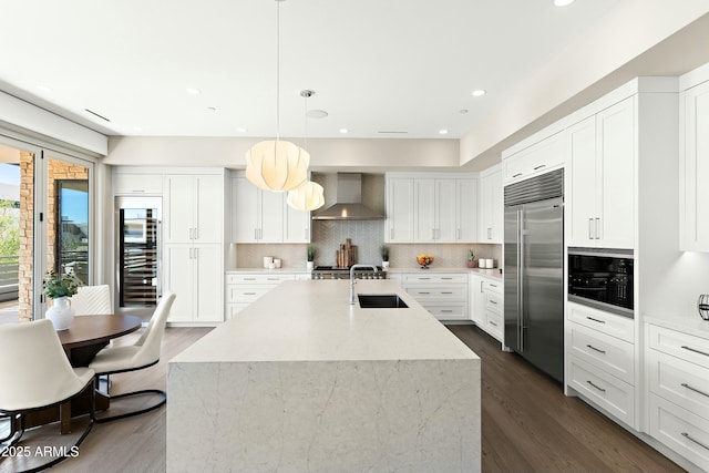 kitchen with built in appliances, beverage cooler, hanging light fixtures, wall chimney range hood, and an island with sink