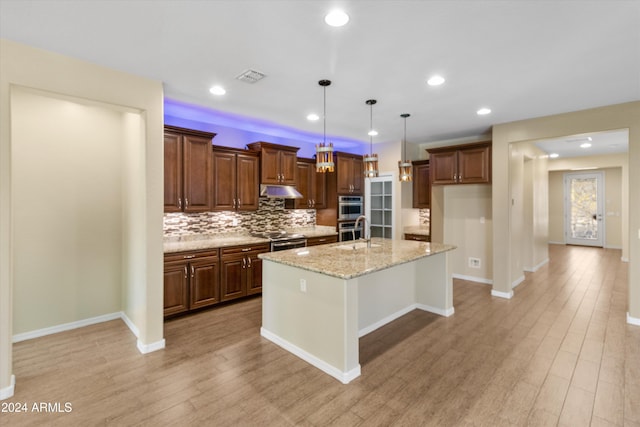 kitchen featuring a kitchen island with sink, stainless steel appliances, light stone countertops, pendant lighting, and light wood-type flooring