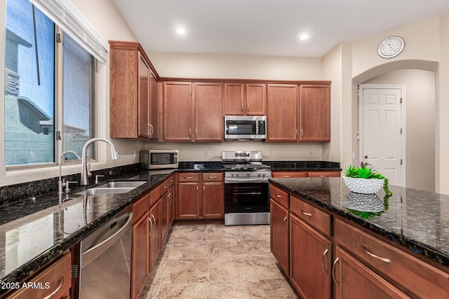 kitchen with appliances with stainless steel finishes, brown cabinets, dark stone countertops, a sink, and recessed lighting