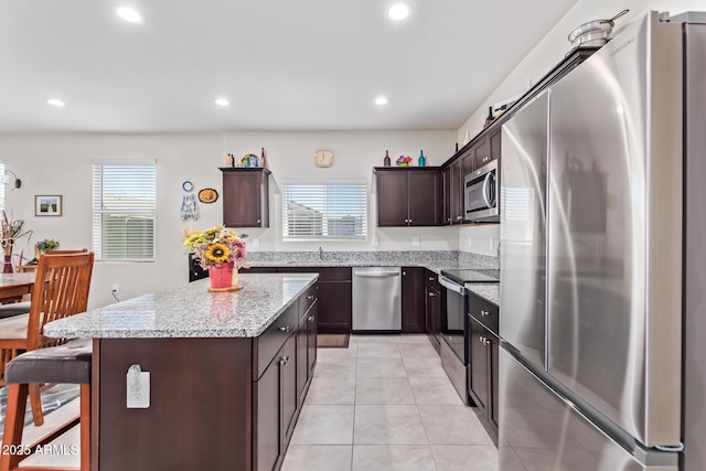 kitchen featuring light tile patterned floors, appliances with stainless steel finishes, a center island, dark brown cabinetry, and light stone countertops