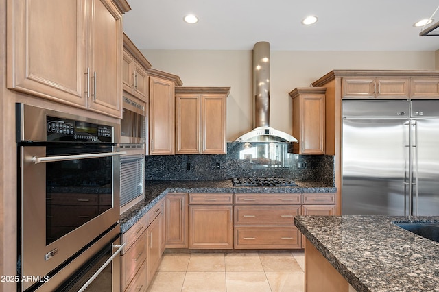 kitchen with light tile patterned floors, stainless steel appliances, tasteful backsplash, dark stone counters, and wall chimney exhaust hood