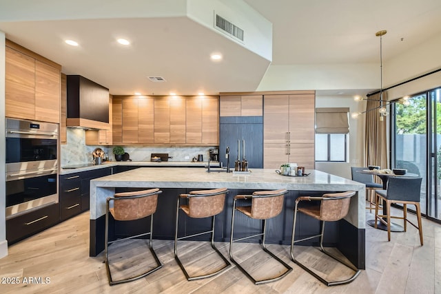 kitchen featuring a breakfast bar, hanging light fixtures, a center island with sink, double oven, and backsplash