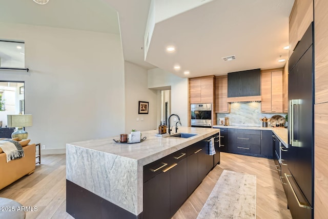 kitchen with light brown cabinetry, sink, a center island with sink, wall chimney range hood, and light hardwood / wood-style flooring