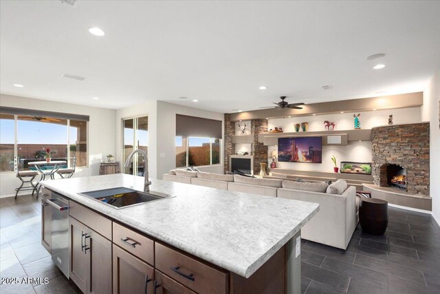 kitchen featuring dishwasher, a stone fireplace, sink, ceiling fan, and an island with sink