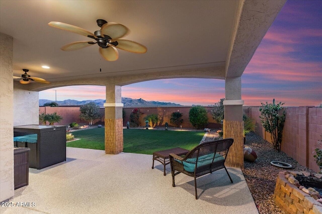 patio terrace at dusk featuring a mountain view, ceiling fan, and a yard