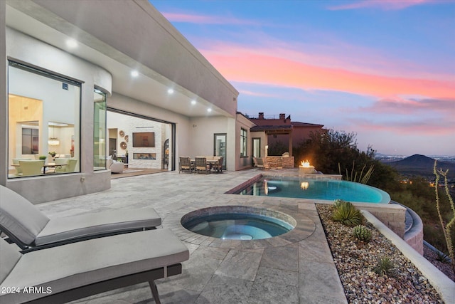 pool at dusk featuring an in ground hot tub, exterior fireplace, and a patio