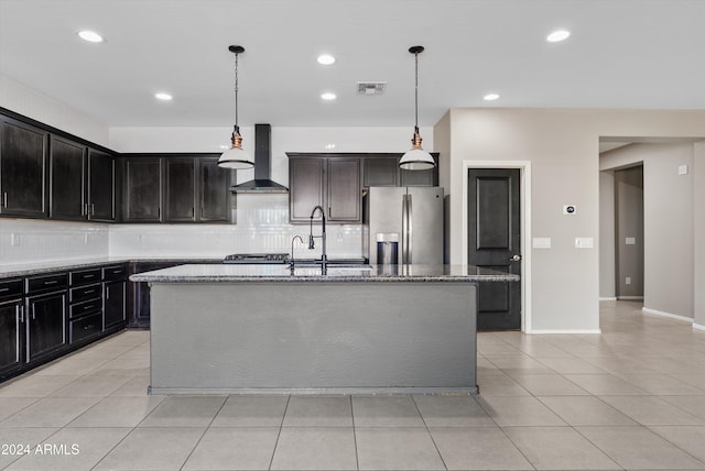 kitchen with wall chimney exhaust hood, hanging light fixtures, light stone counters, stainless steel refrigerator with ice dispenser, and an island with sink