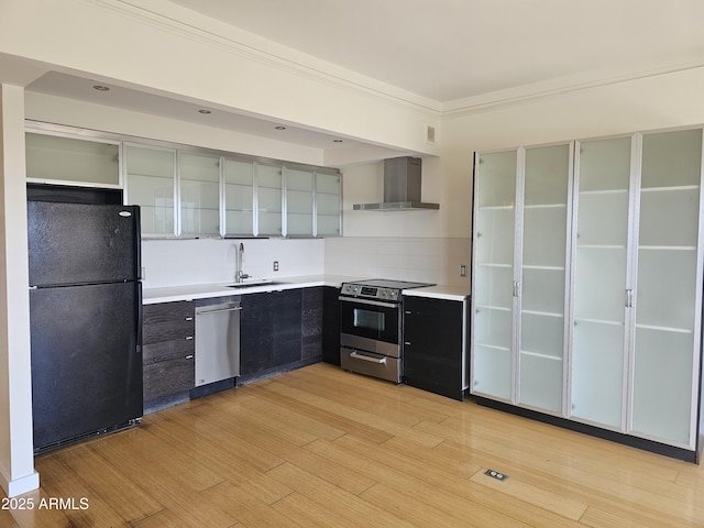 kitchen featuring light wood finished floors, stainless steel appliances, backsplash, wall chimney range hood, and a sink