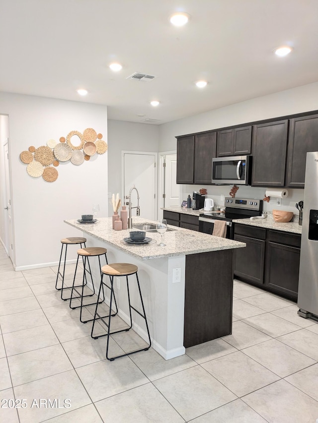 kitchen with appliances with stainless steel finishes, sink, light tile patterned floors, light stone counters, and a center island with sink