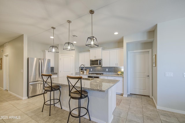 kitchen featuring white cabinets, appliances with stainless steel finishes, a kitchen island with sink, light stone counters, and decorative backsplash