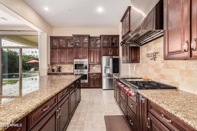 kitchen with light tile patterned flooring, appliances with stainless steel finishes, backsplash, light stone counters, and wall chimney range hood