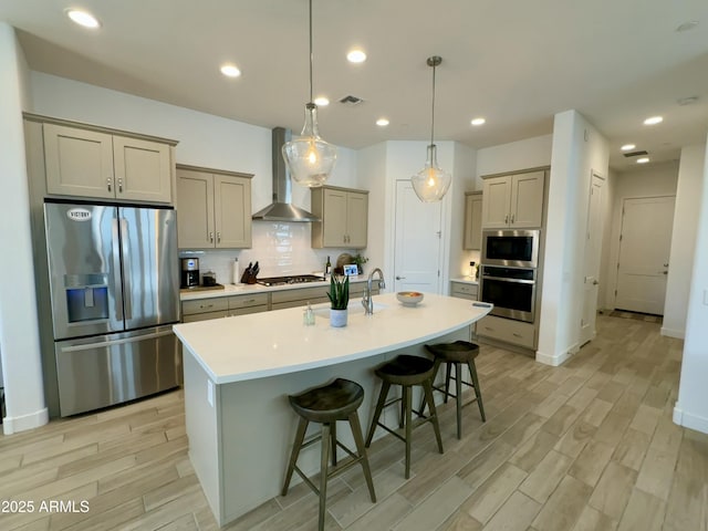 kitchen featuring wall chimney exhaust hood, light wood-style flooring, a kitchen bar, an island with sink, and appliances with stainless steel finishes