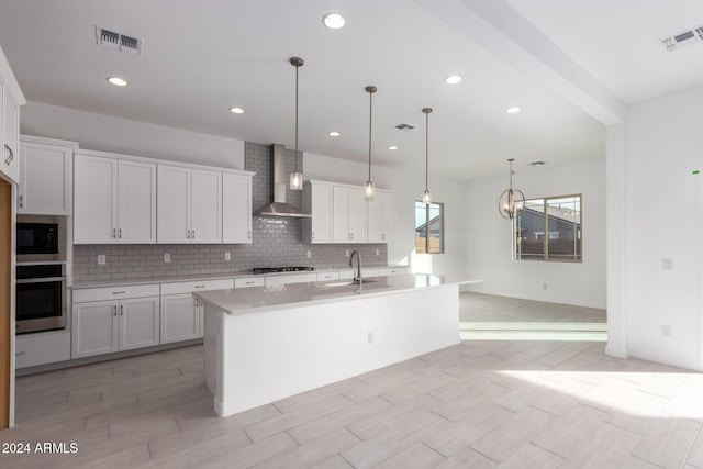 kitchen with hanging light fixtures, white cabinetry, wall chimney range hood, an island with sink, and stainless steel oven