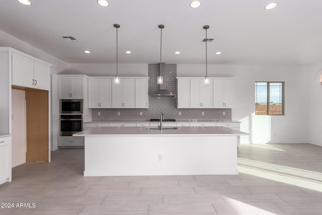 kitchen featuring white cabinets, hanging light fixtures, wall chimney range hood, and a center island with sink