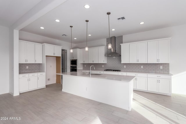 kitchen with wall chimney range hood, decorative light fixtures, white cabinets, sink, and a center island with sink