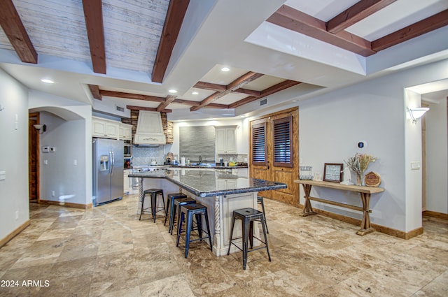 kitchen featuring decorative backsplash, beamed ceiling, a kitchen breakfast bar, custom range hood, and stainless steel refrigerator with ice dispenser