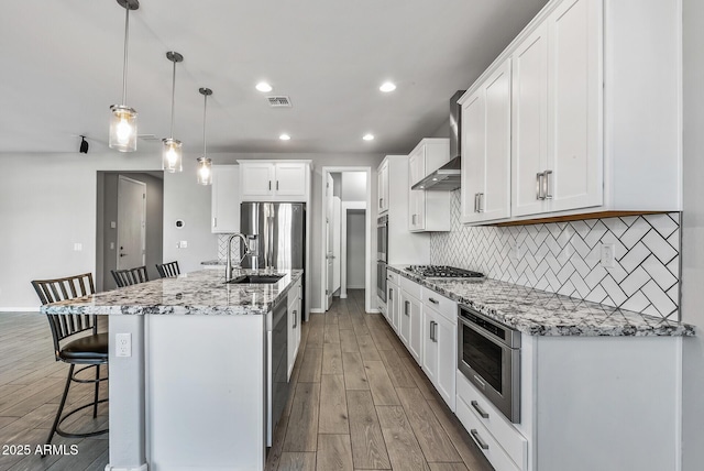 kitchen featuring pendant lighting, wall chimney range hood, a kitchen bar, a kitchen island with sink, and white cabinets