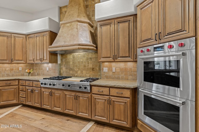 kitchen featuring light hardwood / wood-style flooring, backsplash, stainless steel appliances, premium range hood, and light stone countertops