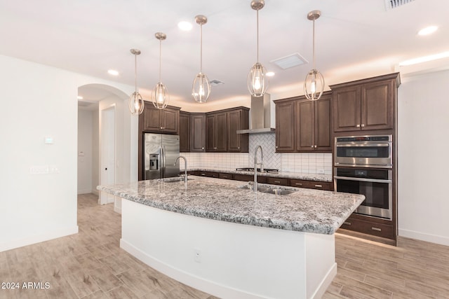 kitchen featuring an island with sink, appliances with stainless steel finishes, wall chimney exhaust hood, and dark brown cabinetry