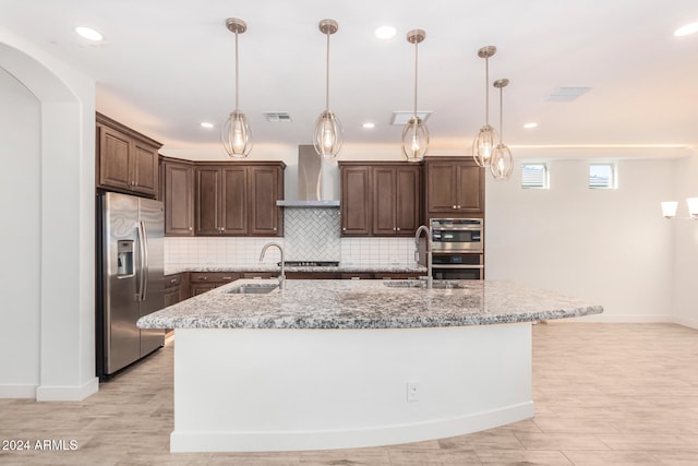 kitchen with wall chimney range hood, sink, a kitchen island with sink, and appliances with stainless steel finishes