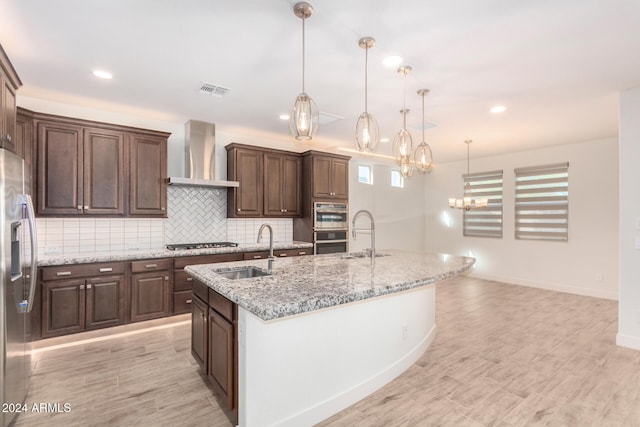 kitchen with a center island with sink, sink, wall chimney exhaust hood, hanging light fixtures, and dark brown cabinets