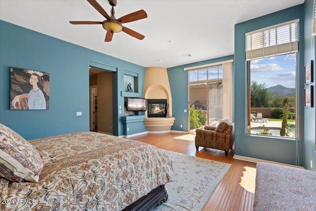 bedroom featuring ceiling fan, a multi sided fireplace, wood-type flooring, and multiple windows