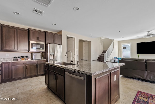 kitchen with appliances with stainless steel finishes, an island with sink, sink, light stone counters, and dark brown cabinets