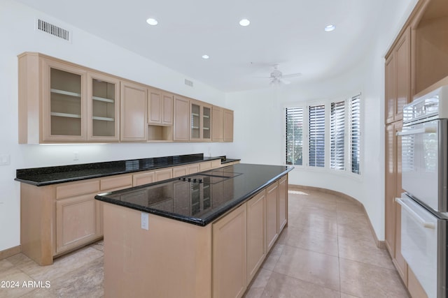 kitchen with light brown cabinetry, dark stone counters, white double oven, ceiling fan, and a kitchen island