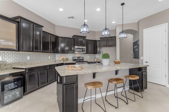 kitchen featuring appliances with stainless steel finishes, backsplash, pendant lighting, a center island with sink, and wine cooler