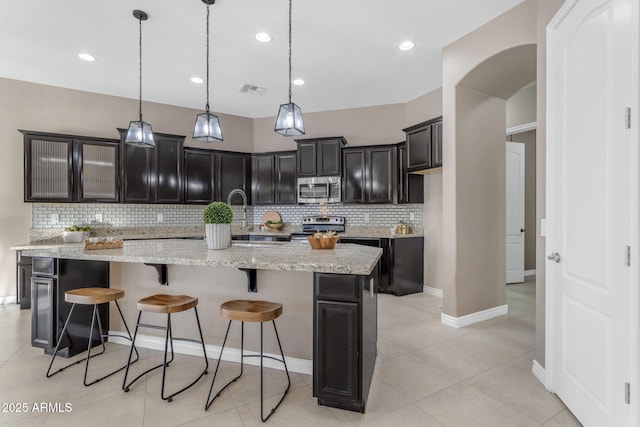 kitchen with light stone countertops, appliances with stainless steel finishes, decorative backsplash, hanging light fixtures, and an island with sink