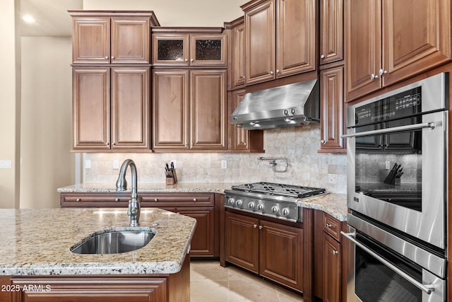 kitchen featuring range hood, light stone countertops, a sink, stainless steel appliances, and backsplash