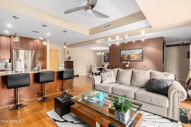 living room featuring ceiling fan with notable chandelier, a tray ceiling, sink, and light hardwood / wood-style flooring