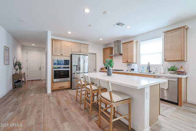 10964 W Garfield St, Avondale, AZ 85323 - kitchen featuring stainless steel appliances, wall chimney exhaust hood, light brown cabinets, a kitchen breakfast bar, and light wood finished floors