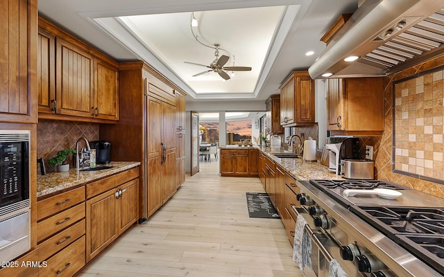 kitchen with high end stainless steel range oven, a tray ceiling, light stone countertops, exhaust hood, and light wood-type flooring