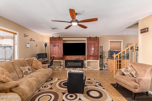 tiled living room with ceiling fan, a barn door, visible vents, stairway, and a glass covered fireplace