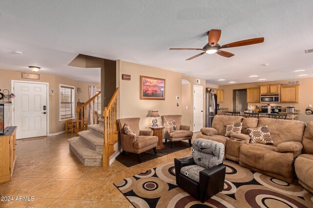 living room with light tile patterned floors, visible vents, baseboards, a ceiling fan, and stairway