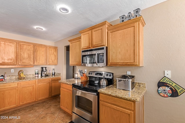 kitchen with a textured ceiling, appliances with stainless steel finishes, light tile patterned floors, and light stone counters