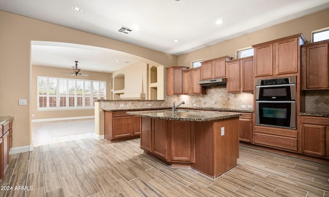 kitchen featuring double oven, backsplash, black electric stovetop, an island with sink, and dark stone counters