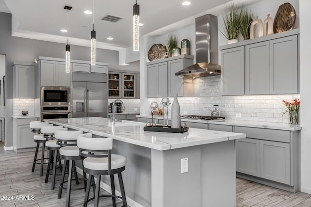 kitchen featuring wall chimney exhaust hood, hanging light fixtures, built in appliances, a spacious island, and light hardwood / wood-style floors