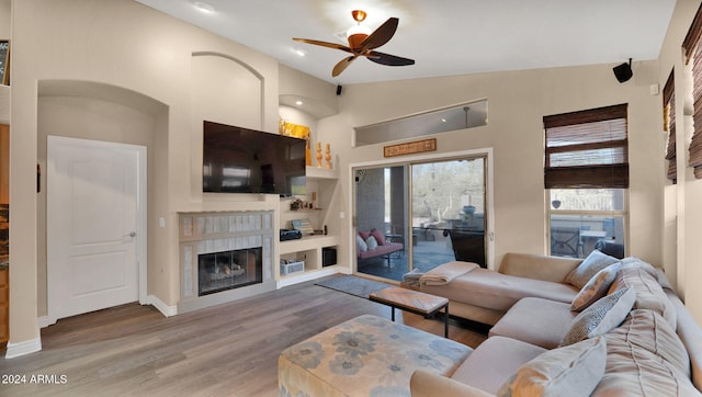 living room with ceiling fan, built in features, vaulted ceiling, a tiled fireplace, and light wood-type flooring