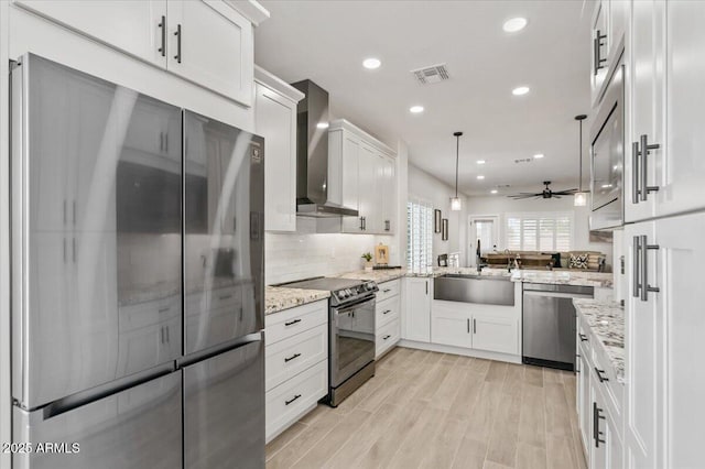 kitchen with appliances with stainless steel finishes, hanging light fixtures, light stone countertops, white cabinets, and wall chimney exhaust hood