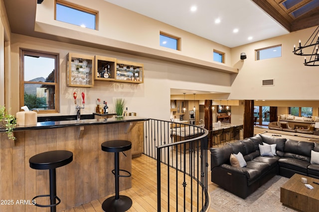 bar with a notable chandelier, visible vents, light wood-style flooring, a high ceiling, and wet bar