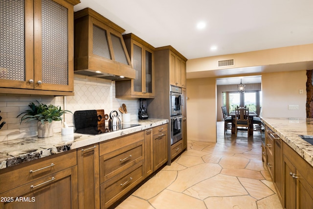 kitchen with black electric stovetop, custom exhaust hood, visible vents, decorative backsplash, and brown cabinetry