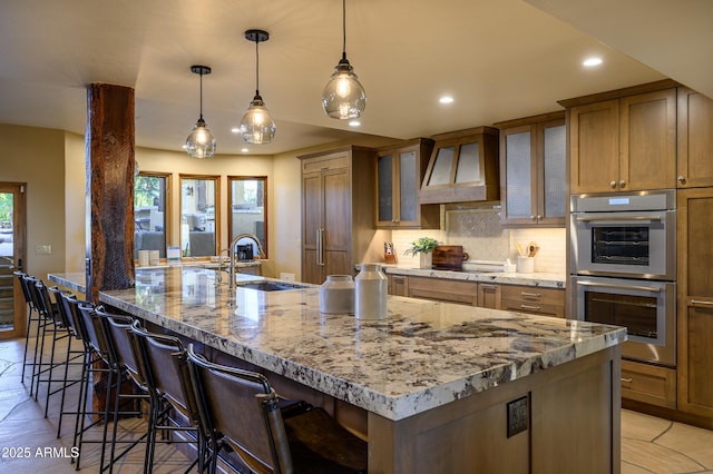 kitchen featuring light stone counters, backsplash, stainless steel double oven, a sink, and premium range hood