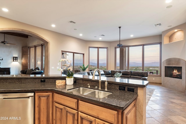 kitchen featuring ceiling fan, a tiled fireplace, dark stone counters, stainless steel dishwasher, and sink