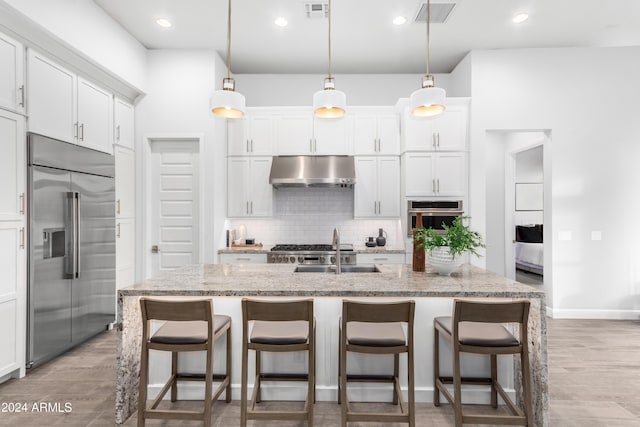 kitchen featuring ventilation hood, an island with sink, light stone countertops, and appliances with stainless steel finishes