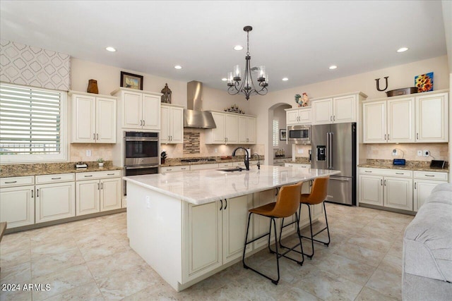 kitchen with sink, wall chimney range hood, hanging light fixtures, appliances with stainless steel finishes, and a center island with sink