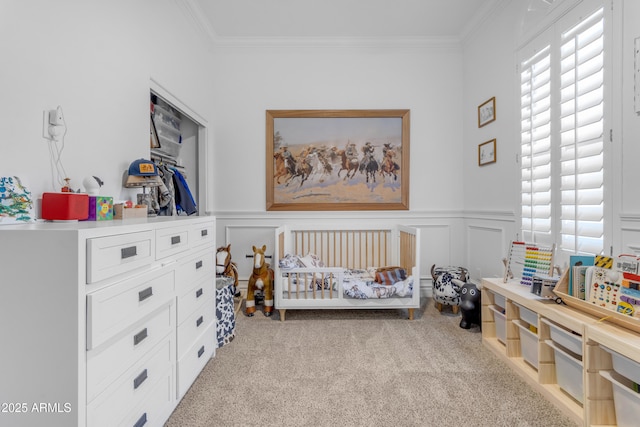 carpeted bedroom featuring a crib, a decorative wall, wainscoting, and crown molding