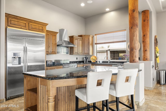 kitchen featuring wall chimney range hood, a breakfast bar, dark stone countertops, stainless steel appliances, and a center island