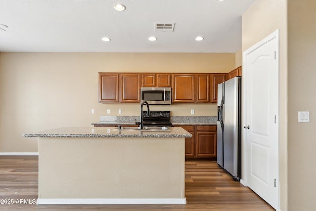 kitchen featuring appliances with stainless steel finishes, a sink, wood finished floors, brown cabinets, and a center island with sink
